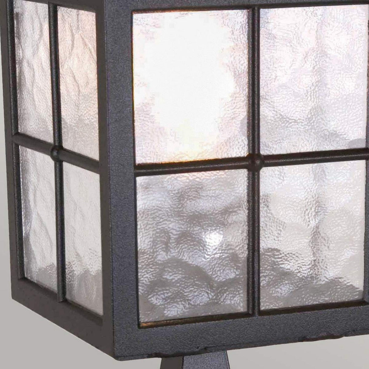 Close-up of the Winchester 1 Light Pedestal Light - Black, featuring a rectangular design made with textured glass panes and a die-cast aluminum frame. The glowing light inside is gently diffused through frosted glass, creating an inviting glow that exudes vintage charm while mounted on a metal stand.