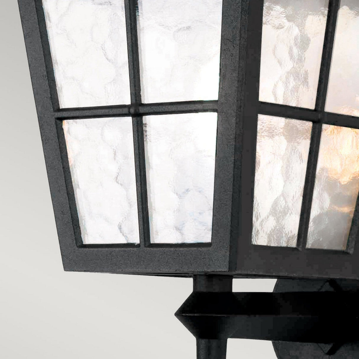 Close-up of the Canterbury Outdoor Wall Lantern Up in black, crafted from die-cast aluminium. It features a modern design with textured glass panels and grid-like frames. The warm glow of the interior light contrasts beautifully against a neutral gray background, seamlessly blending contemporary style with traditional charm.