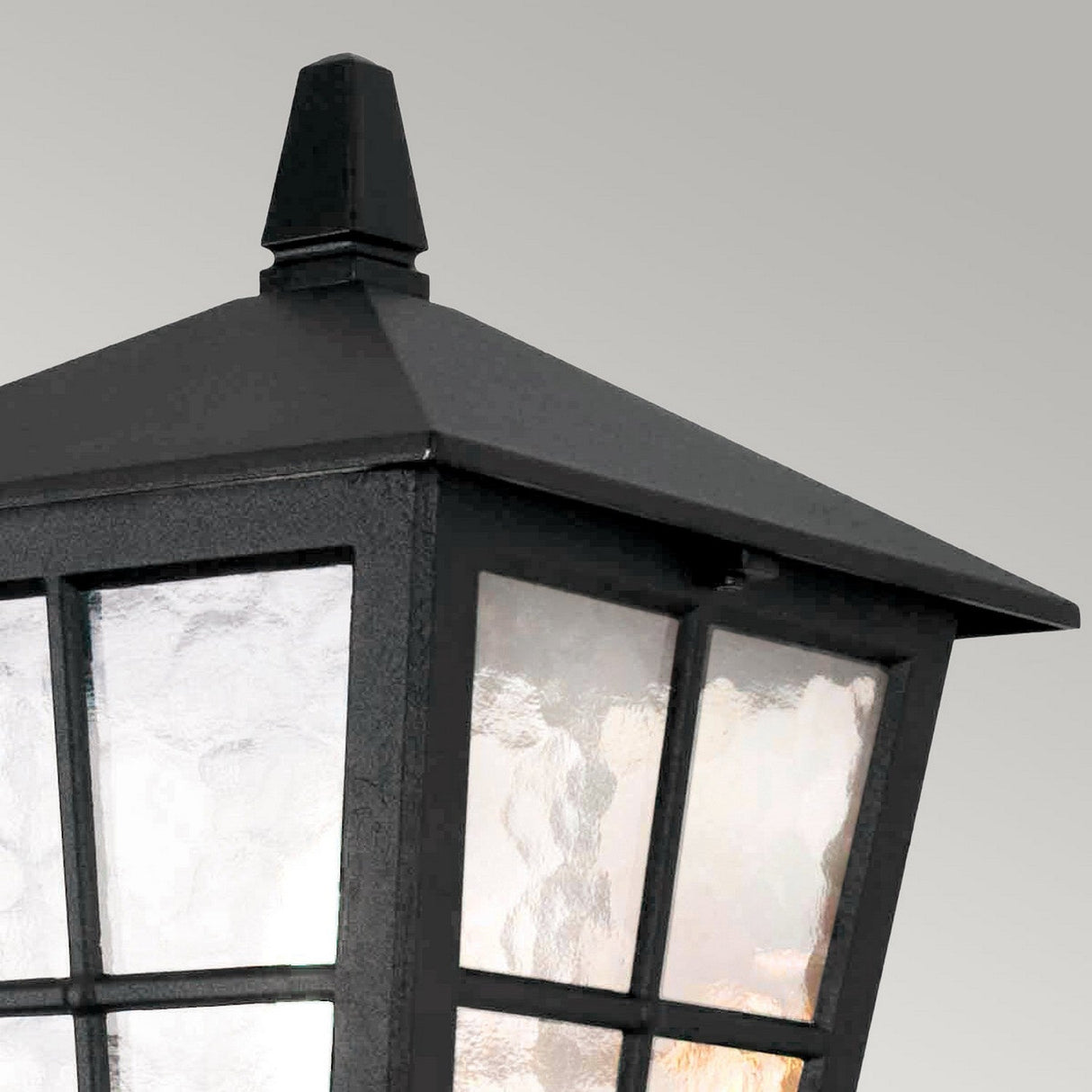 Close-up of a Canterbury Outdoor Wall Lantern Up - Black, crafted from die-cast aluminium. It features frosted glass panes and a pointed top, set against a light gray background. The traditional design emits a soft glow that suggests it is illuminated.