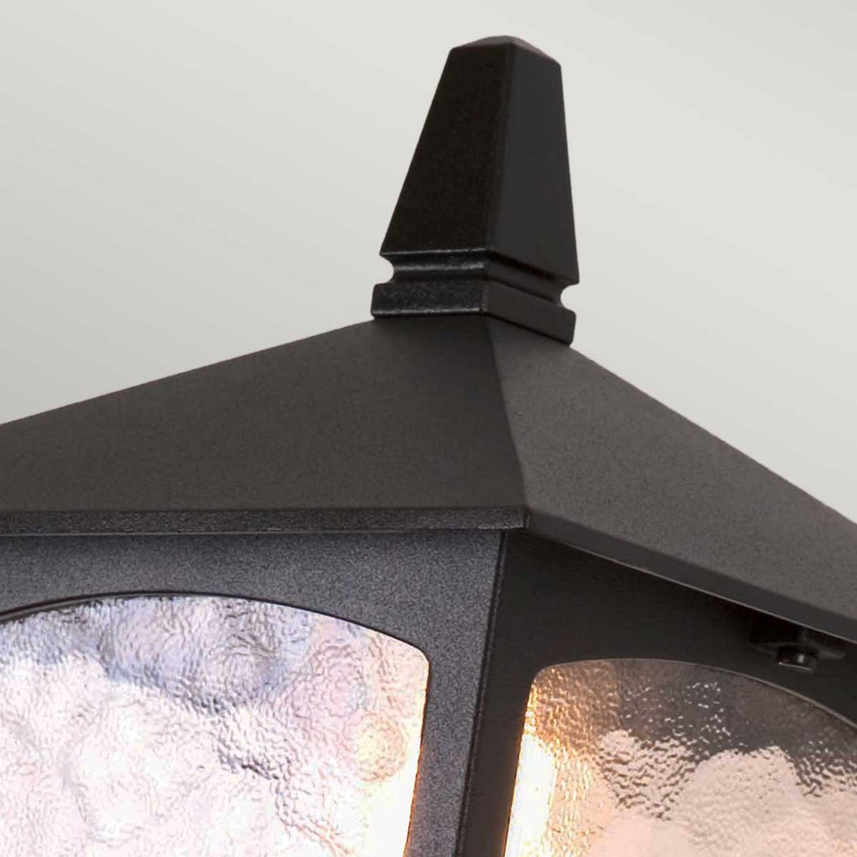 Close-up of the York 1 Light Outdoor Wall Up Light in black, featuring a pyramid-shaped top and textured glass panels. This weather-resistant fixture emits a warm glow through the glass, exuding vintage-inspired lighting appeal. The background is a soft, light gray.