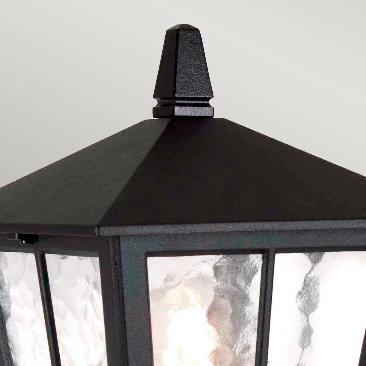 Close-up of the Canterbury Outdoor Pedestal Lantern - Black, showcasing its black metal frame and pyramid-shaped top with textured glass panels. This British design lights up from within, emitting a gentle glow. Its robust construction ensures enduring elegance against the plain, light gray background.