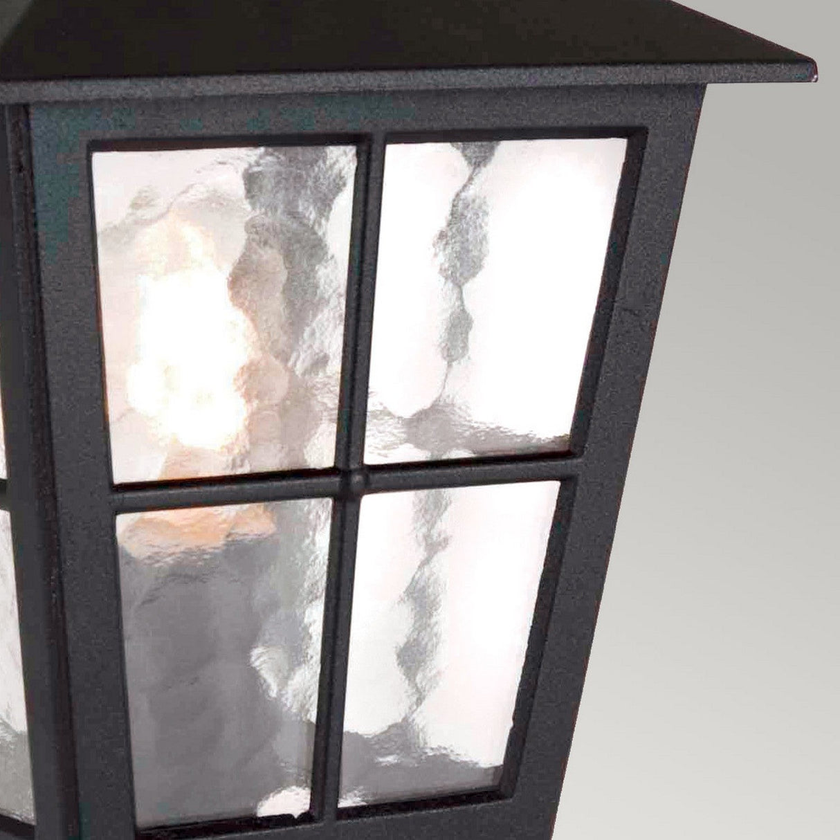 Close-up of the Canterbury Outdoor Pedestal Lantern - Black, featuring its sturdy build with black-framed textured glass panels and a gentle, glowing light within. The design draws inspiration from classic British aesthetics and is set against a simple, neutral background, accentuating its elegance.