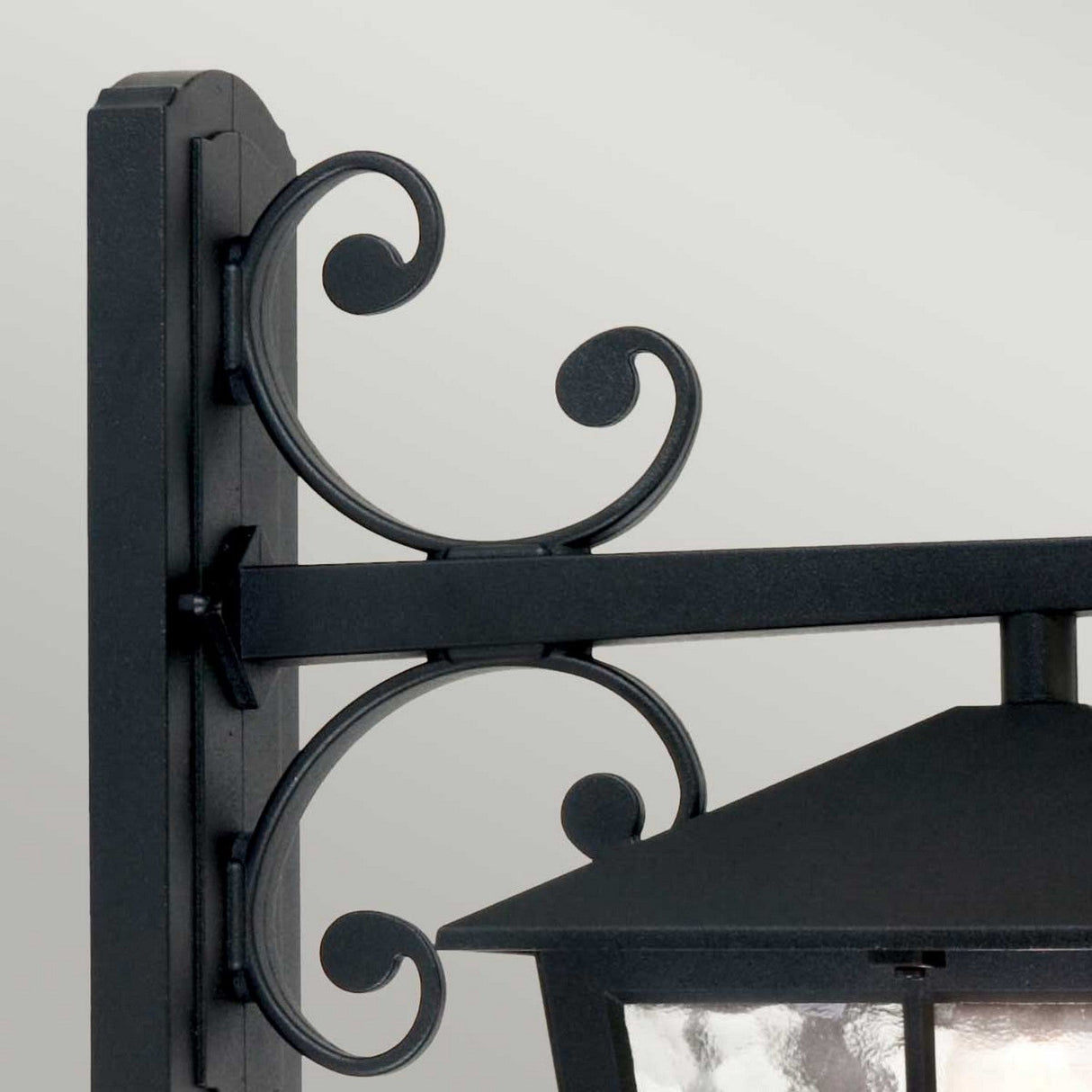 Close-up of the Canterbury Outdoor Wall Lantern Down in black, mounted on a wall. This traditional lighting piece showcases decorative scrollwork and a glass enclosure, offering a classic and elegant appearance. Crafted from durable die-cast aluminum, it promises lasting beauty in any setting.