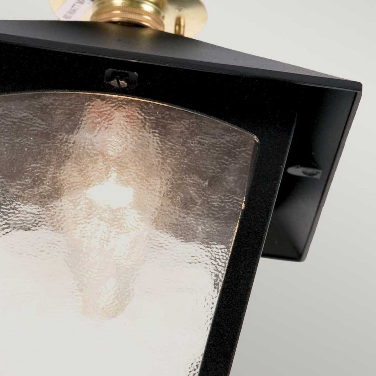 Close-up view of the York 1 Light Outdoor Flush Porch Light - Black, featuring a rectangular design with a black die-cast aluminium frame and transparent textured glass, showcasing a glowing E27 bulb inside. The fixture is mounted at an angle on a ceiling.