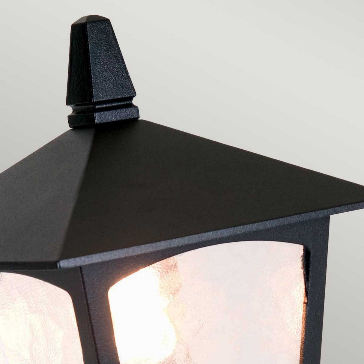 A close-up of the York 1 Light Pedestal Light - Black, showcasing its die-cast aluminum design and vintage charm with a pyramid-shaped top. It emits a soft glow that accentuates the textured glass panels against a pale background.