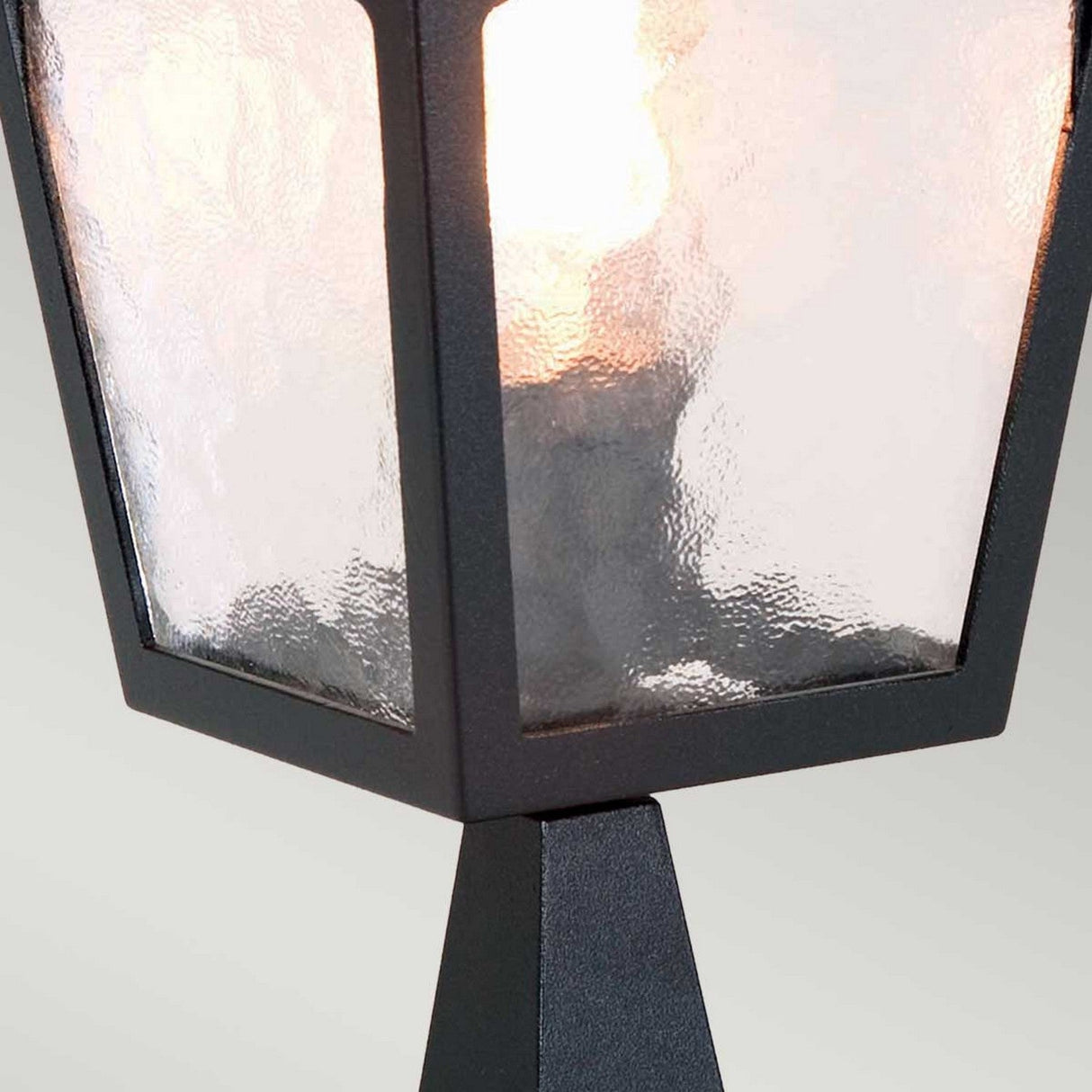 Close-up of a York 1 Light Pedestal Light - Black, showcasing vintage charm with its black design and frosted glass panels. The gently glowing light enhances outdoor illumination, creating a warm and inviting ambiance against the plain, light-colored background.