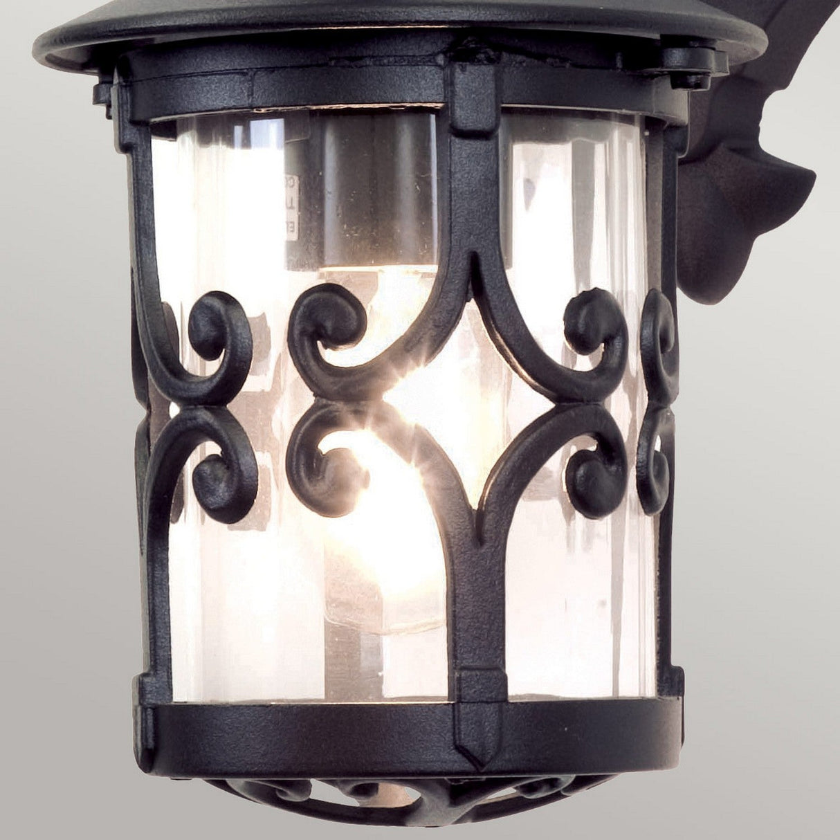 The Hereford Outdoor Wall Down Lantern - Black is a lantern-style fixture made from die-cast aluminum, featuring detailed scrollwork and clear glass panels that enclose a glowing light bulb against a plain background.