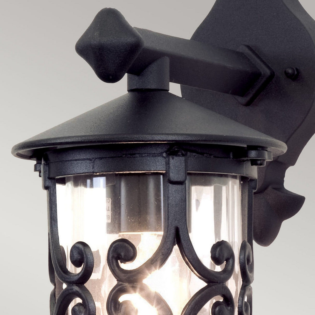 The Hereford Outdoor Wall Down Lantern - Black is a decorative fixture styled like a lantern, featuring clear glass casing and intricate metal scrollwork. Crafted from die-cast aluminium, it is mounted on a wall bracket with a simple and elegant design for timeless appeal.