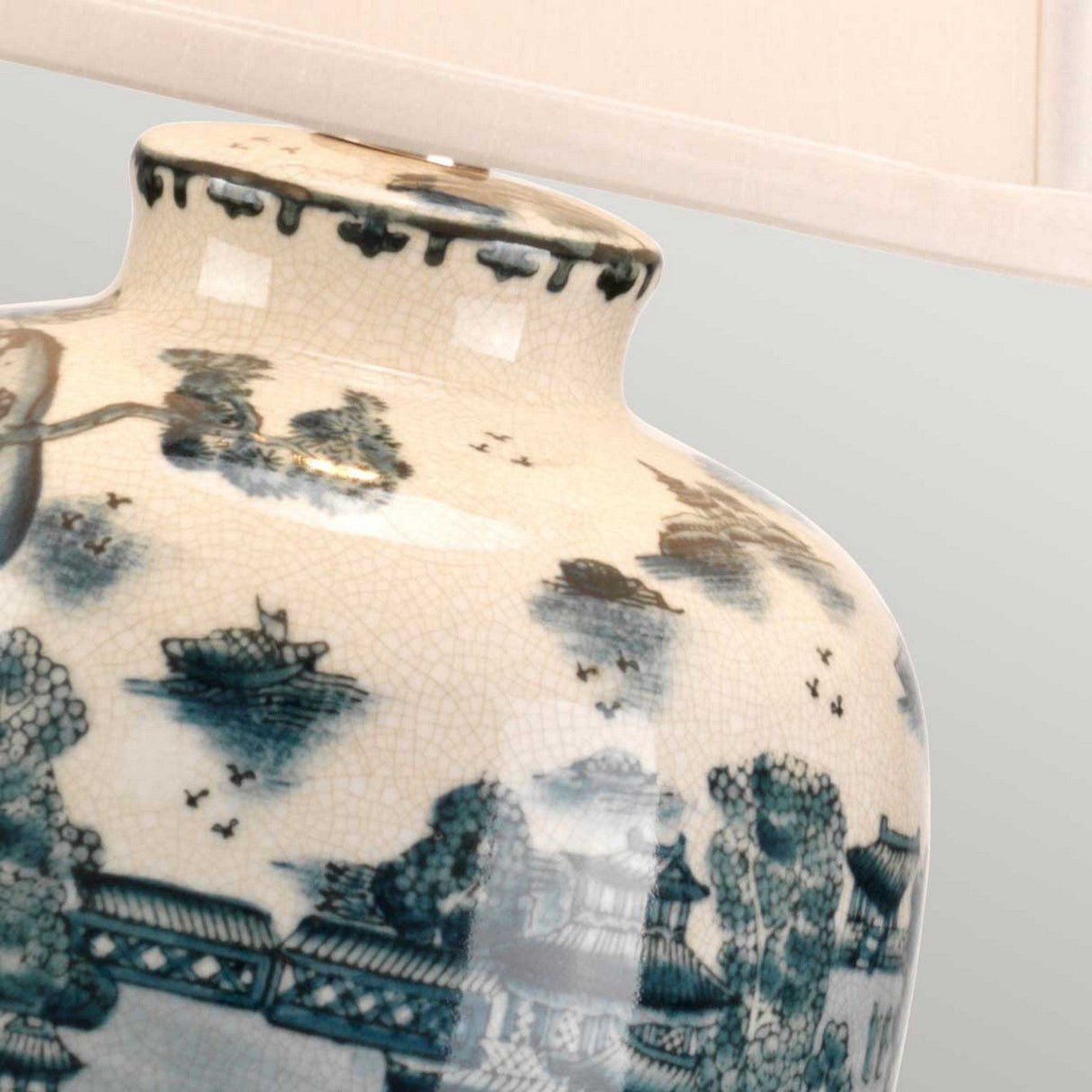 A detailed view of the Blue Traditional Table Lamp - Blue & White showcases its decorative ceramic base adorned with intricate blue and white designs reminiscent of Chinese porcelain. It beautifully depicts traditional Asian landscapes, complete with pagodas and trees, enhancing the lamp's cultural charm against a light background. The top portion also reveals a cream-colored lampshade.