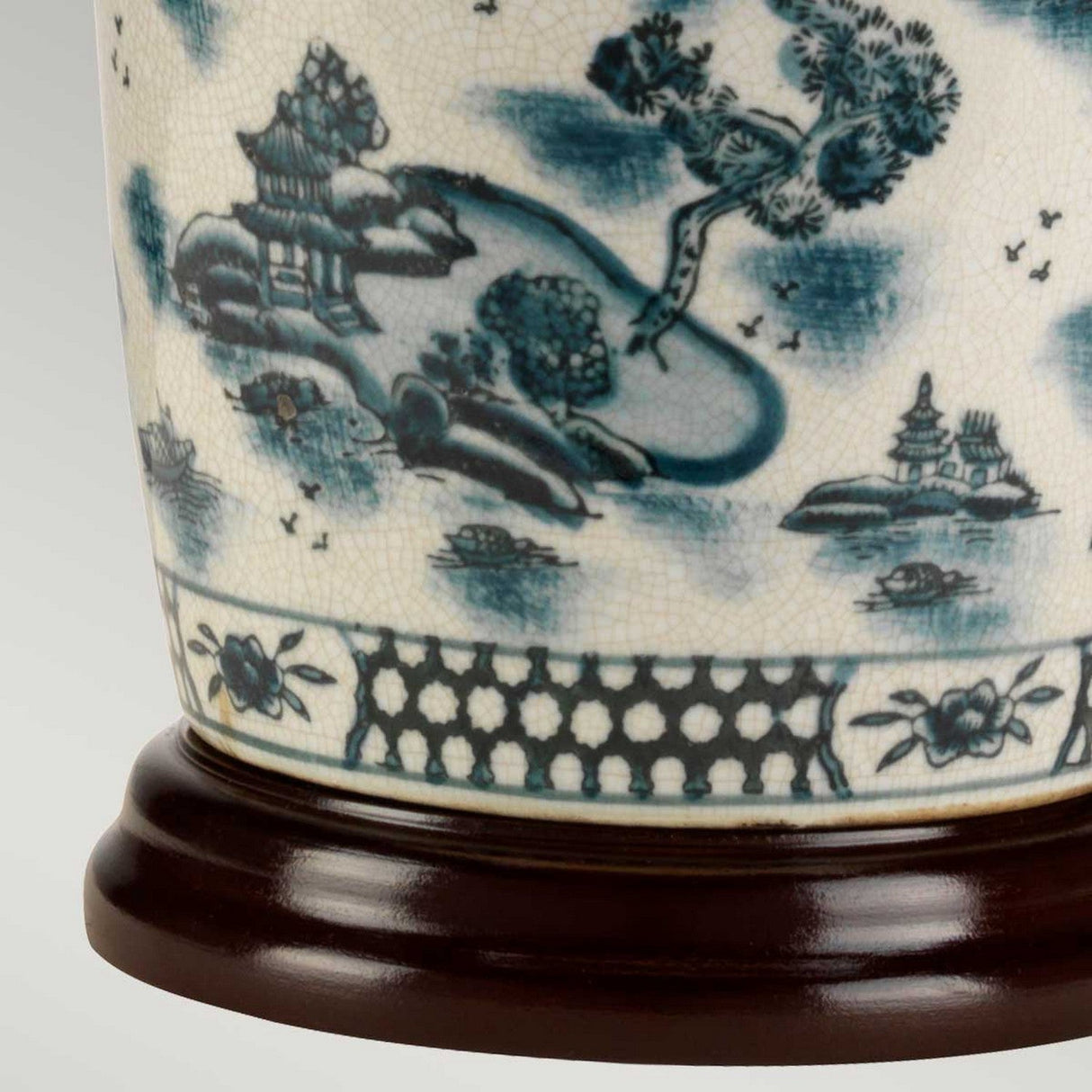 Close-up of a Chinese porcelain vase with a blue and white landscape design featuring trees, a building, and rocks. The vase exudes cultural charm as it rests on a dark wooden base adorned with floral patterns and geometric shapes, reminiscent of the Blue Traditional Table Lamp - Blue & White.