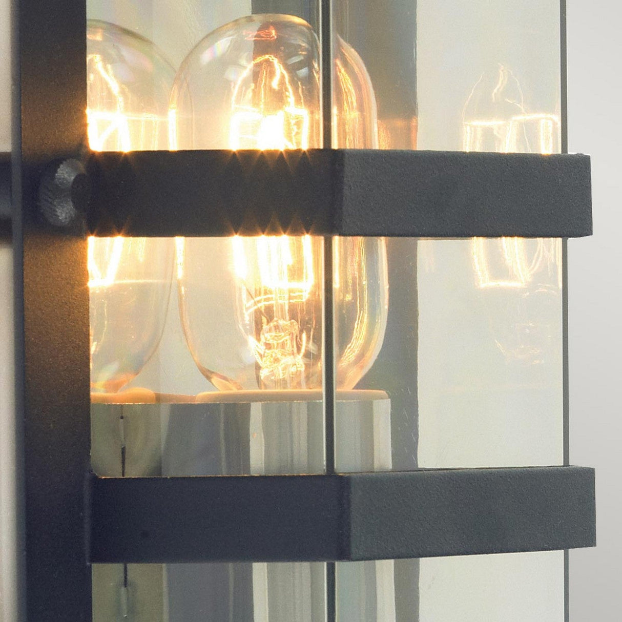 A close-up of the Boden Outdoor 1 Light Wall Light - Black With Clear Glass reveals two glowing filament bulbs within a clear glass enclosure, framed by black metal bands in an art deco design, with the warm light softly diffusing through the glass.