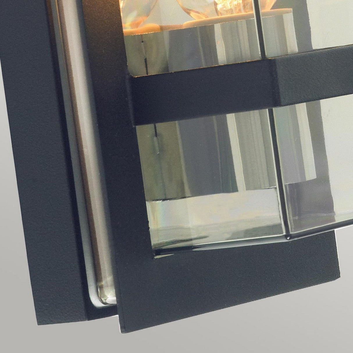 A close-up of the Boden Outdoor 1 Light Wall Light, featuring a sleek Art Deco design with a black metal frame and clear glass panels. The light is switched on, casting a warm glow through the transparent sections, ideal for outdoor use thanks to its IP54 rating.