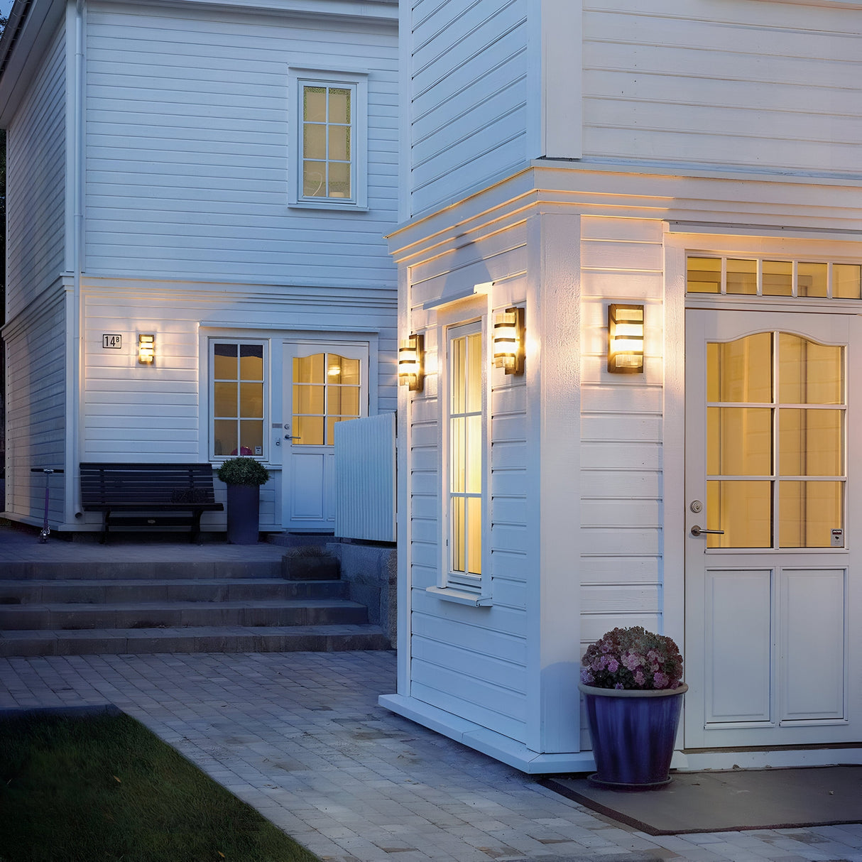 At dusk, a cozy, modern home exterior is highlighted by its white wooden walls and warm indoor lighting. The entrance features a clear glass door complemented by black Boden Outdoor 1 Light Wall Lights with clear glass, which showcase an Art Deco design. A bench and potted plant are placed to the left, near steps that lead up to the inviting doorway.