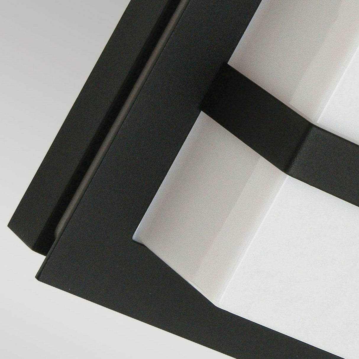 Close-up of the Boden Outdoor 1 Light Wall Light in black with frosted glass, showcasing its modern geometric design. The black frame is angled above the frosted glass fixture, creating a sleek, minimalist appearance against a light gray background.