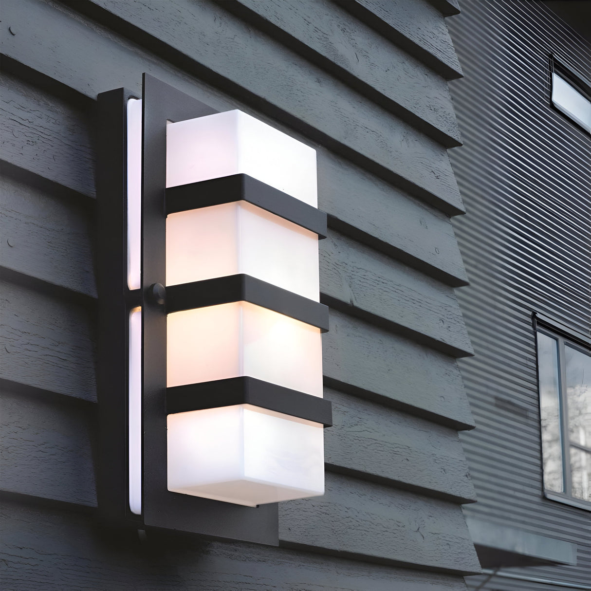 The Boden Outdoor 1 Light Wall Light in black with frosted glass is mounted on a dark-toned exterior wall, featuring a modern Art Deco rectangular design with black horizontal accents. It emits a soft, warm glow, while a window with reflective glass appears in the background.