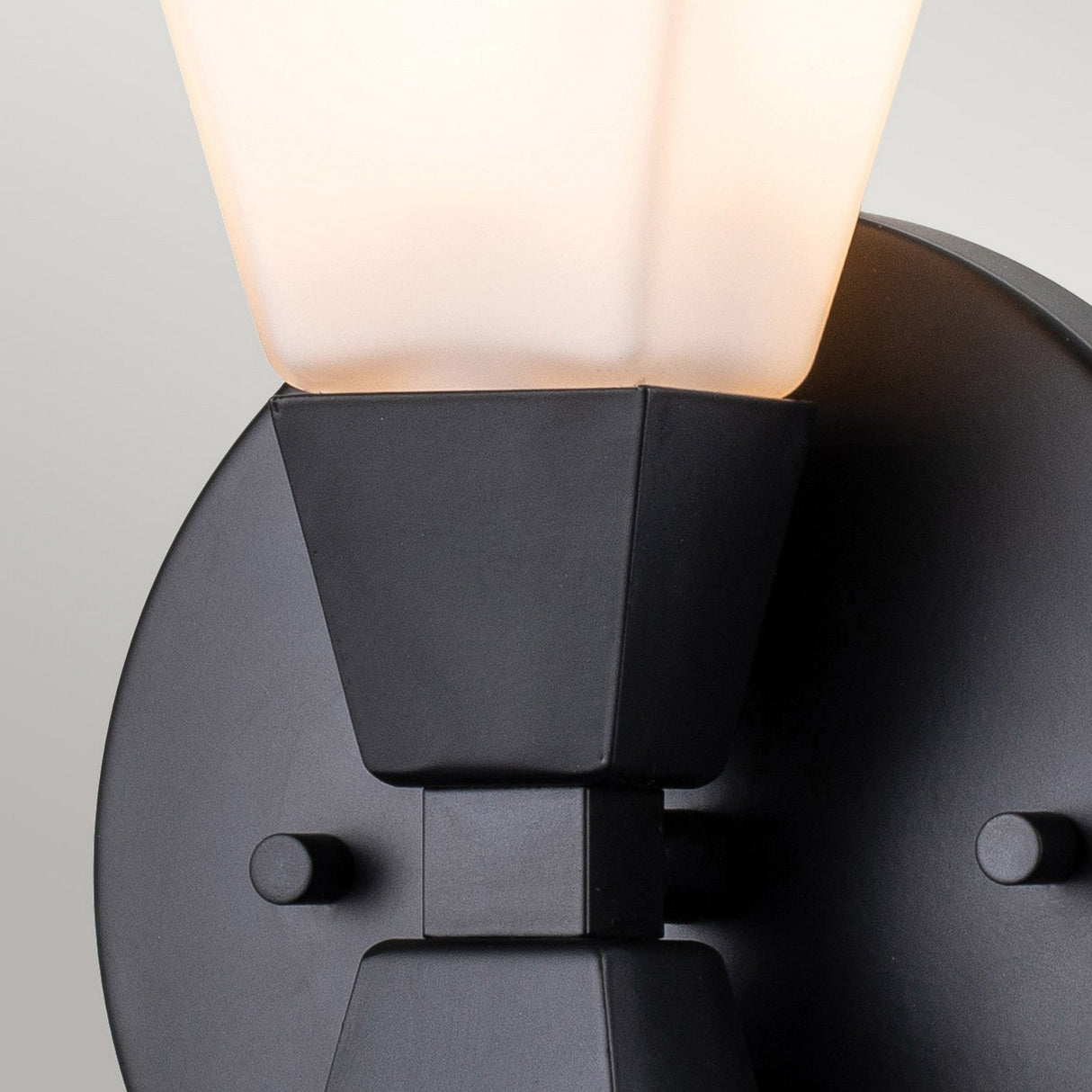 Close-up of the Bowtie 1 Light Wall Light in a modern matte black finish, showcasing its minimalistic design. The fixture includes a circular base and a frosted, geometric-shaped glass shade, softly illuminating against a light gray background—ideal for stylish bathroom lighting.