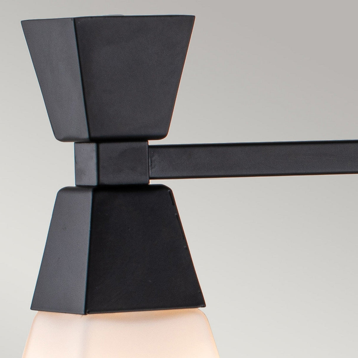 A close-up of the Bowtie 3 Light Wall Light - Matte Black reveals its minimalist design, showcasing a matte black geometric support and rod that holds a white frosted glass shade. The fixture, which is IP44 rated for damp areas, stands out against the neutral gray background.