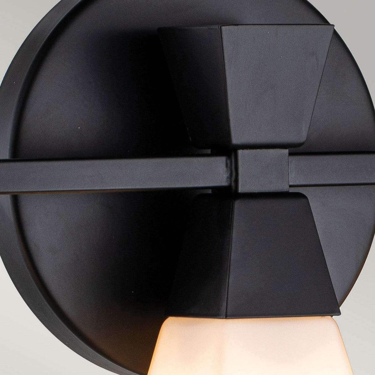 A detailed view of the Bowtie 3 Light Wall Light - Matte Black highlights its contemporary geometric design, with a circular base and triangular light shade that casts a warm glow. This IP44-rated fixture, sleek and minimalist against a light gray backdrop, enhances any space with elegance.