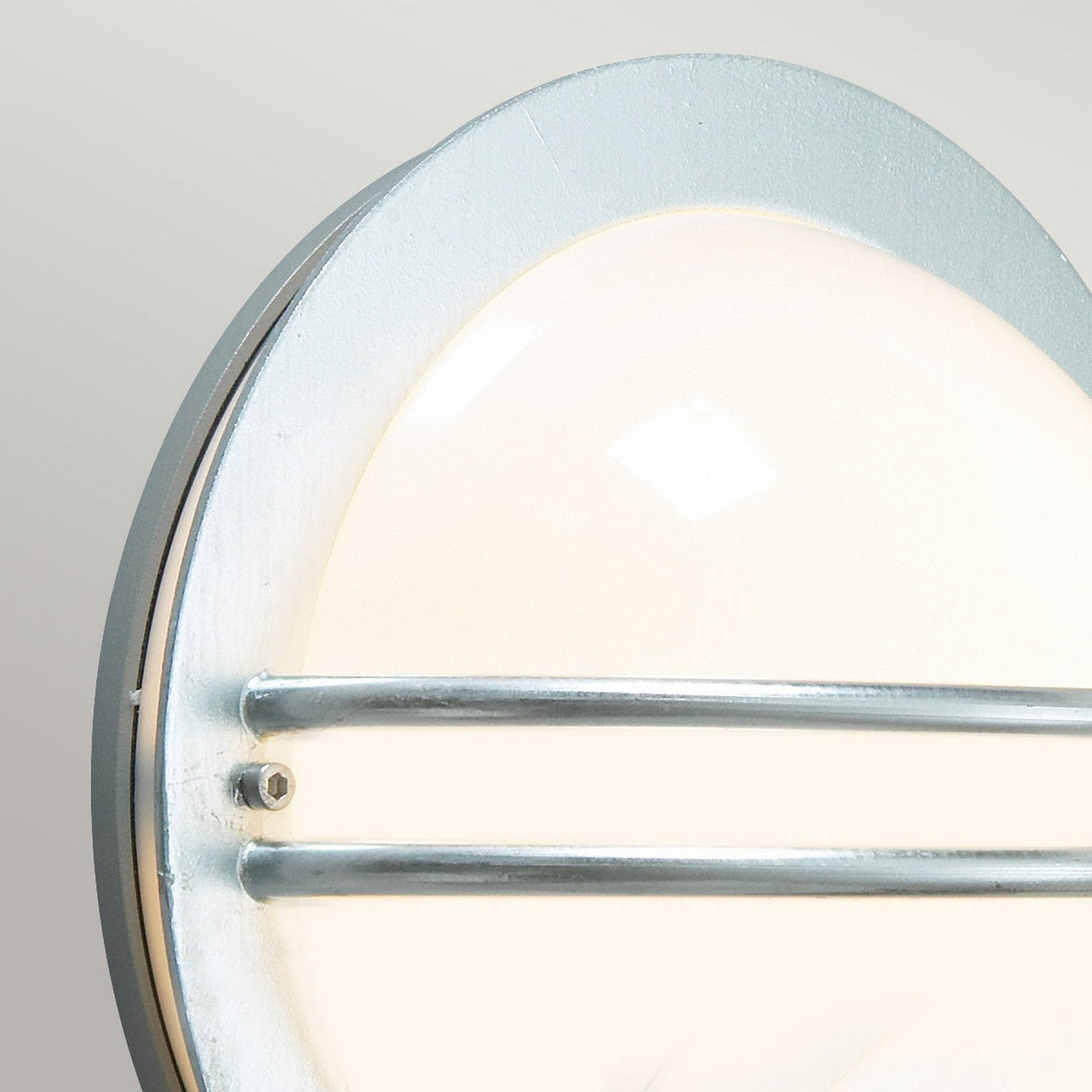 Featuring a close-up of the Bremen Outdoor 1 Light Wall Lantern in silver, this round, modern wall lamp showcases a frosted white cover and galvanized steel frame, exuding a maritime aesthetic. With two horizontal metal bars, it emits a soft, warm glow against a light gray background.