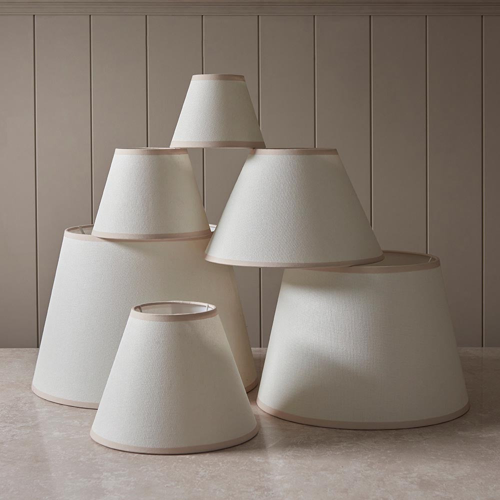 Brockbury tapered lamp shade in vintage white fabric with almond taped edge, shown among various sizes against a neutral panelled backdrop.