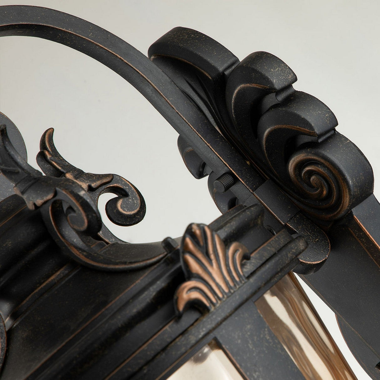 Close-up of the Baltimore 1 Light Outdoor Large Wall Lantern in weathered bronze, showcasing ornate black detailing and curving metalwork with an amber-tinted glass pane. Its intricate design features scroll patterns and a fleur-de-lis motif on the frame, making it ideal as an outdoor wall light.