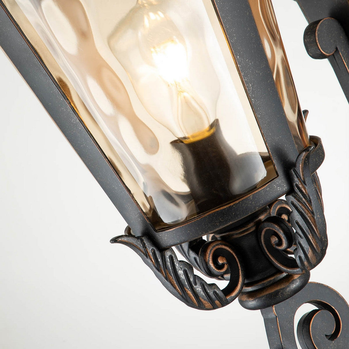 Close-up of the Baltimore 1 Light Outdoor Large Wall Lantern, showcasing its vintage-style features with amber-tinted glass and intricate metal scrollwork. The light bulb inside casts a warm glow, highlighting the elegant accents of its weathered bronze finish.