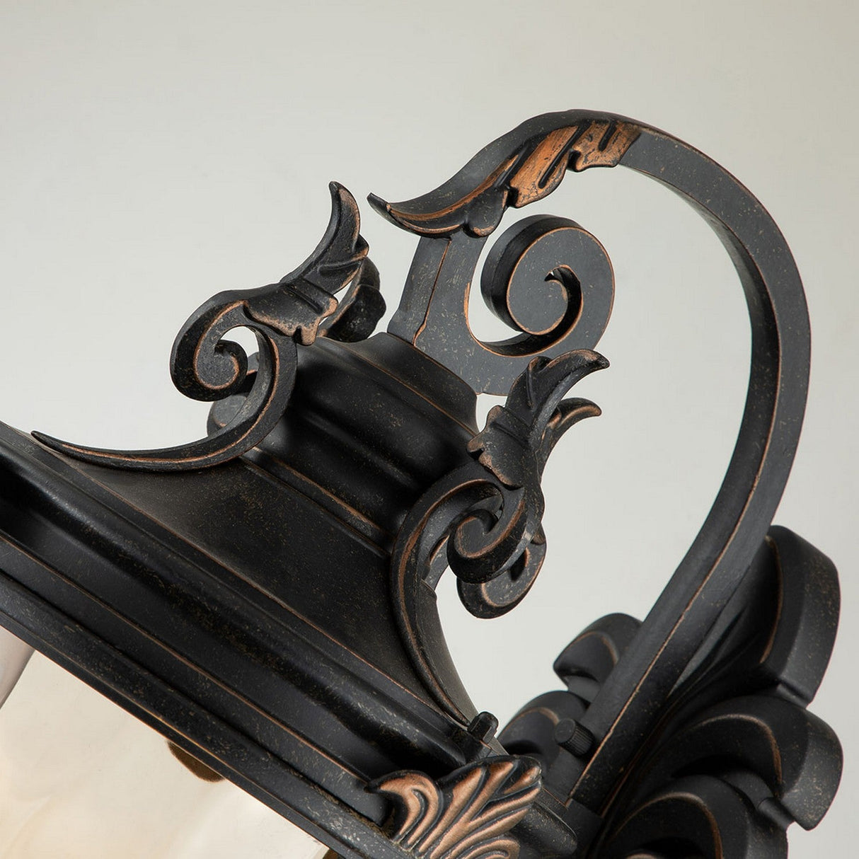 A close-up view of the Baltimore 1 Light Outdoor Large Wall Lantern in weathered bronze showcases its intricate scrollwork and leaf designs. The curved handle contributes an elegant touch to the overall design. Set against a neutral background, the lantern is beautifully enhanced by amber-tinted glass.