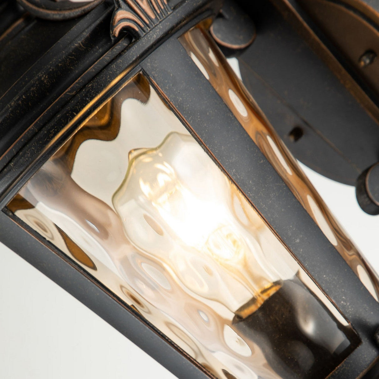 Close-up of the Baltimore 1 Light Outdoor Medium Wall Lantern - Weathered Bronze, showcasing a glowing bulb inside. The lantern's textured glass panels and metal frame enhance its vintage-inspired design, producing a warm, inviting glow.