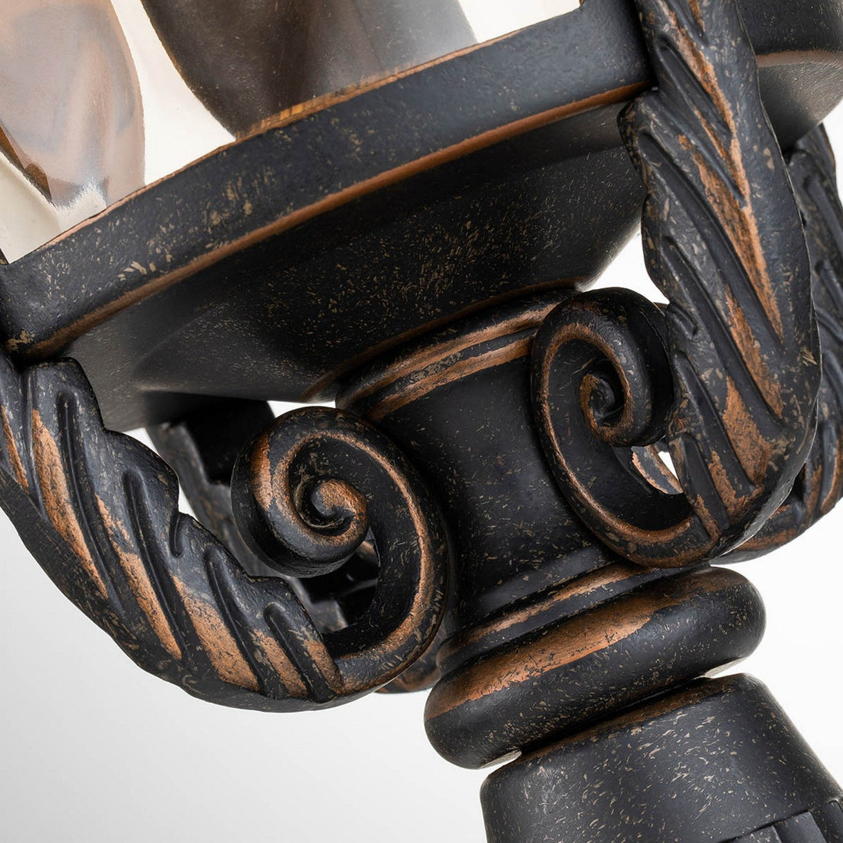 A close-up of the Baltimore 1 Light Large Pedestal Lantern showcases its ornate, weathered bronze-finished metal with intricate scroll and rope-like designs. Its surface exudes a timeworn charm, reminiscent of traditional outdoor lanterns, highlighting the craftsmanship and detailed textures against a light background.