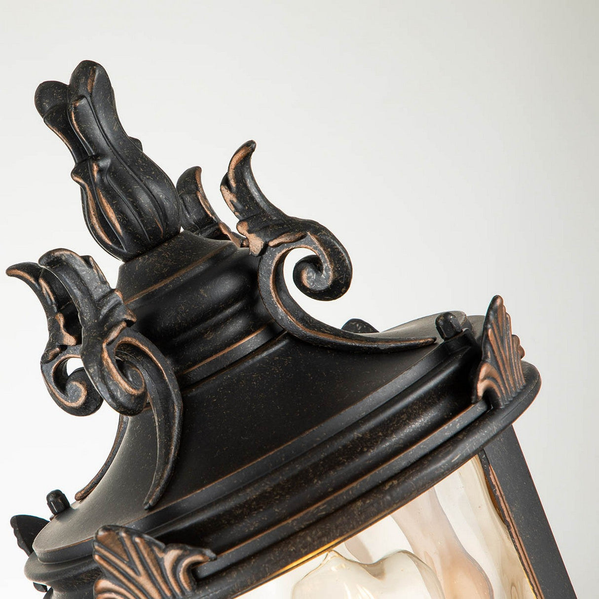 A close-up of the "Baltimore 1 Light Medium Pedestal Lantern - Weathered Bronze" reveals an outdoor lantern crafted from black metal with elegant bronze accents. The design showcases amber-tinted glass and intricate scrollwork, all perched atop a weathered bronze pedestal with a gracefully curved top, exemplifying a timeless and sophisticated style.