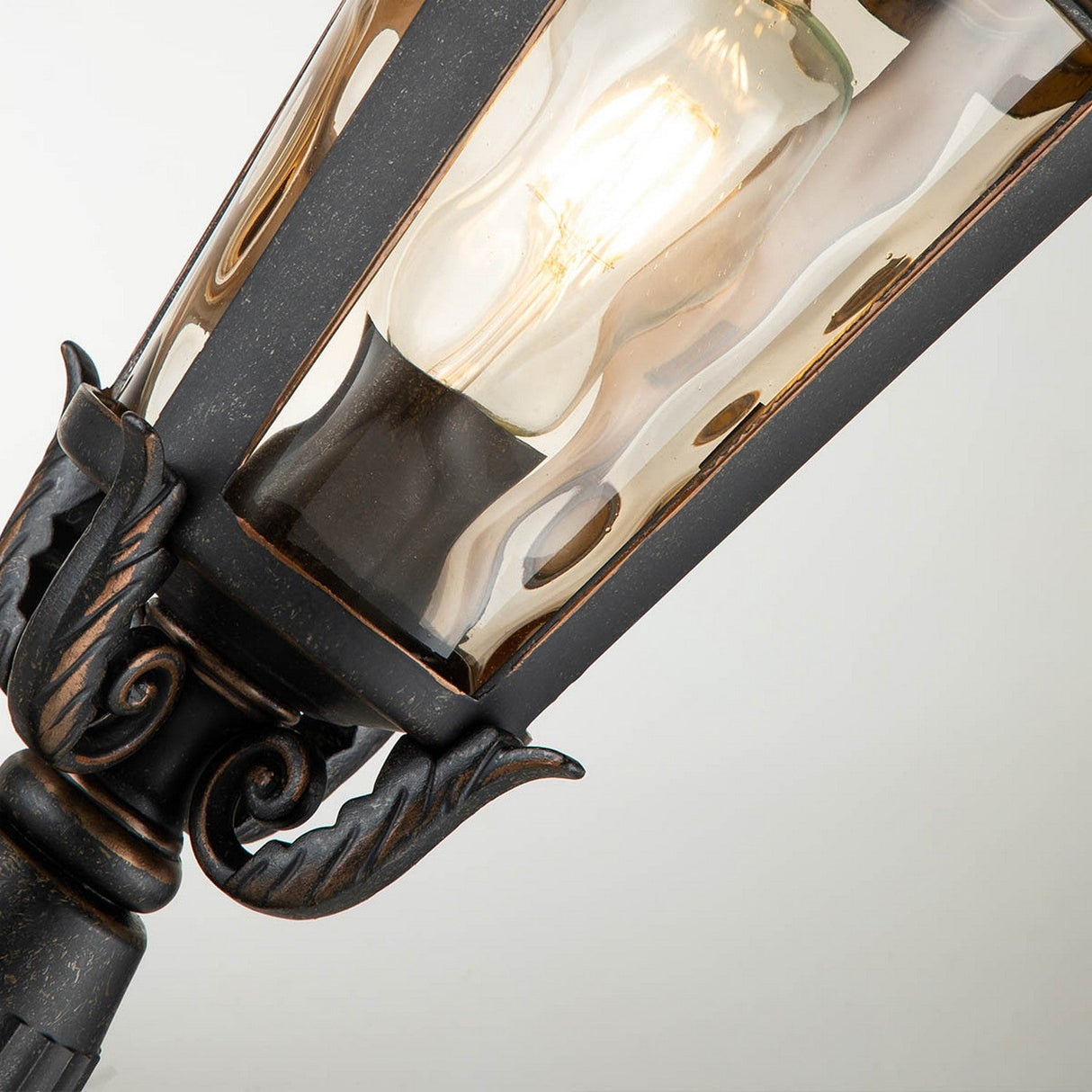 A close-up of the Baltimore 1 Light Medium Pedestal Lantern showcases its ornate design, featuring a weathered bronze pedestal and amber-tinted glass. The lantern is adorned with curved decorative elements and a glass casing that houses a lit bulb, emitting a warm glow.