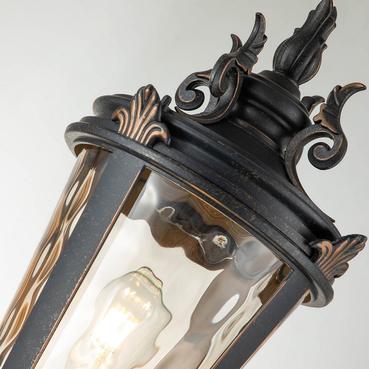 A close-up of the Baltimore 1 Light Medium Pillar showcases its vintage design. This outdoor lighting piece has a black finish with weathered bronze accents and a clear glass cover that reveals a glowing light bulb inside.