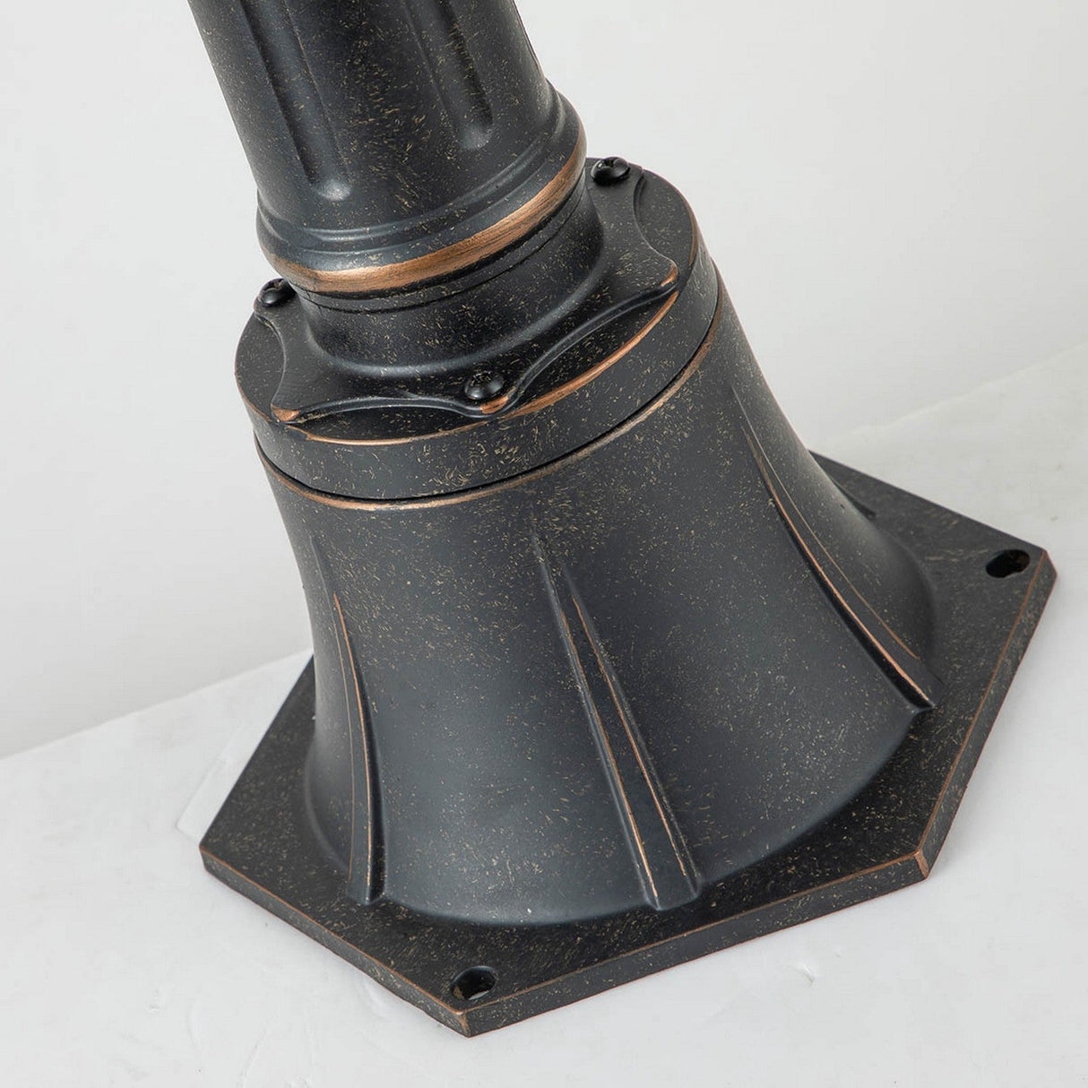 Close-up of the Baltimore 1 Light Medium Pillar in weathered bronze, featuring an ornate base with intricate ridges and bolts, all set against a plain white background.