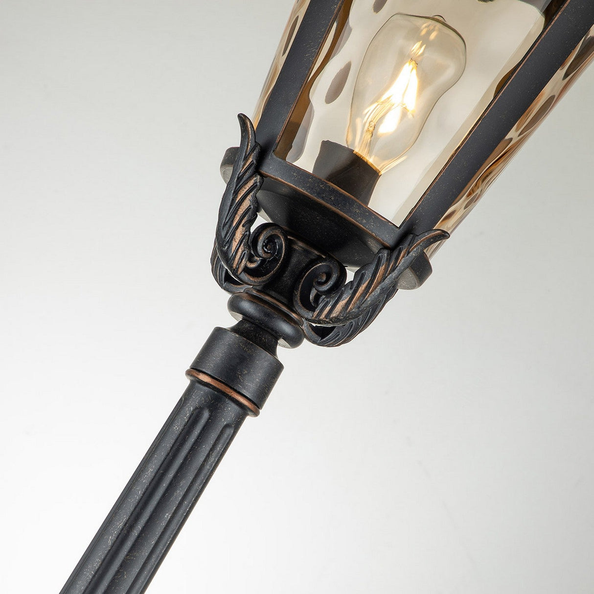 Close-up of the Baltimore 1 Light Large Lamp Post, featuring ornate metal detailing with a clear bulb inside an amber-tinted blown glass enclosure. The intricate scrollwork is accentuated by the Weathered Bronze Patina, standing elegantly against a neutral background.