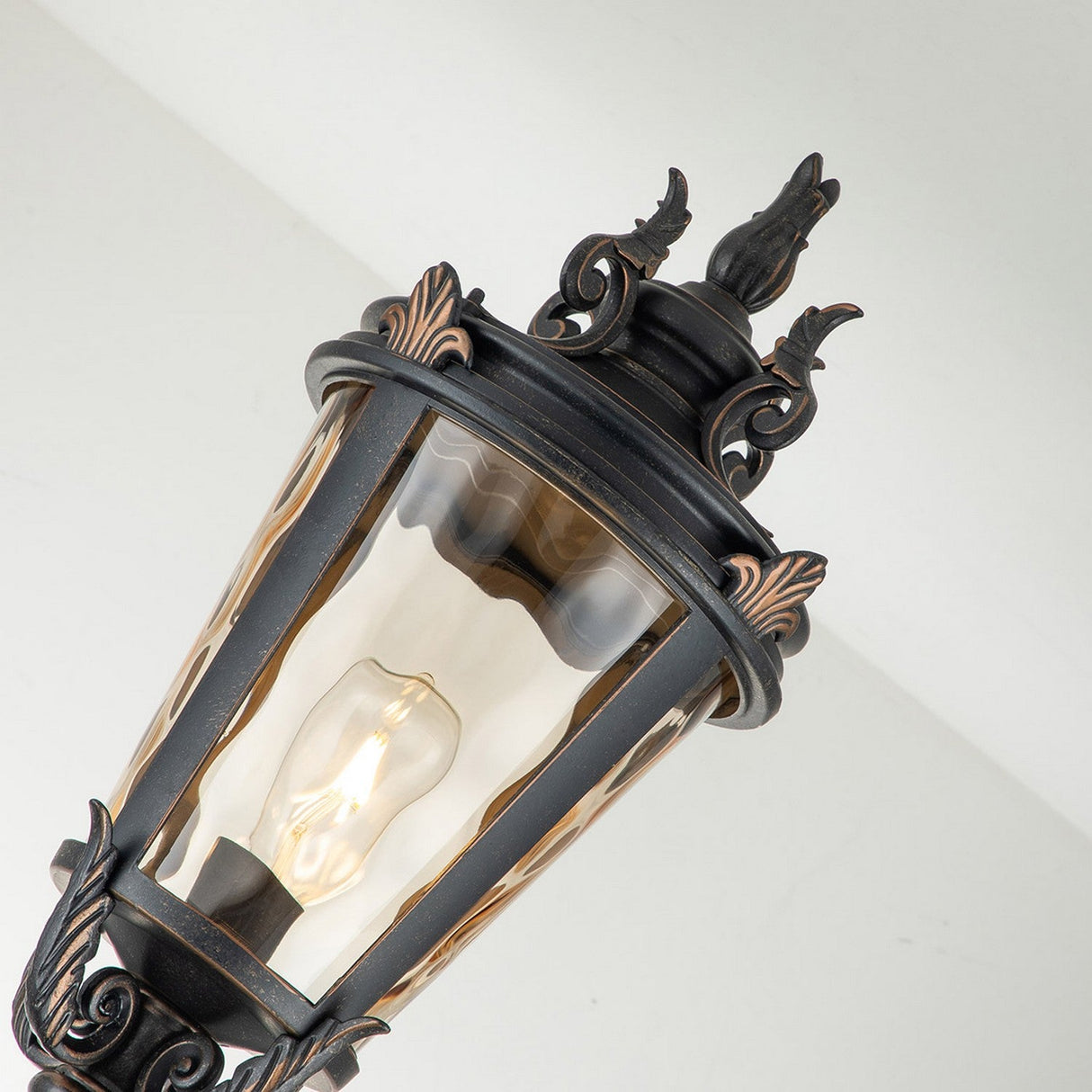 Baltimore 1 Light Large Lamp Post in a weathered bronze finish features an ornate black design with amber-tinted blown glass casing, revealing a visible light bulb. The lamp is displayed against a white wall, accentuating its intricate details.