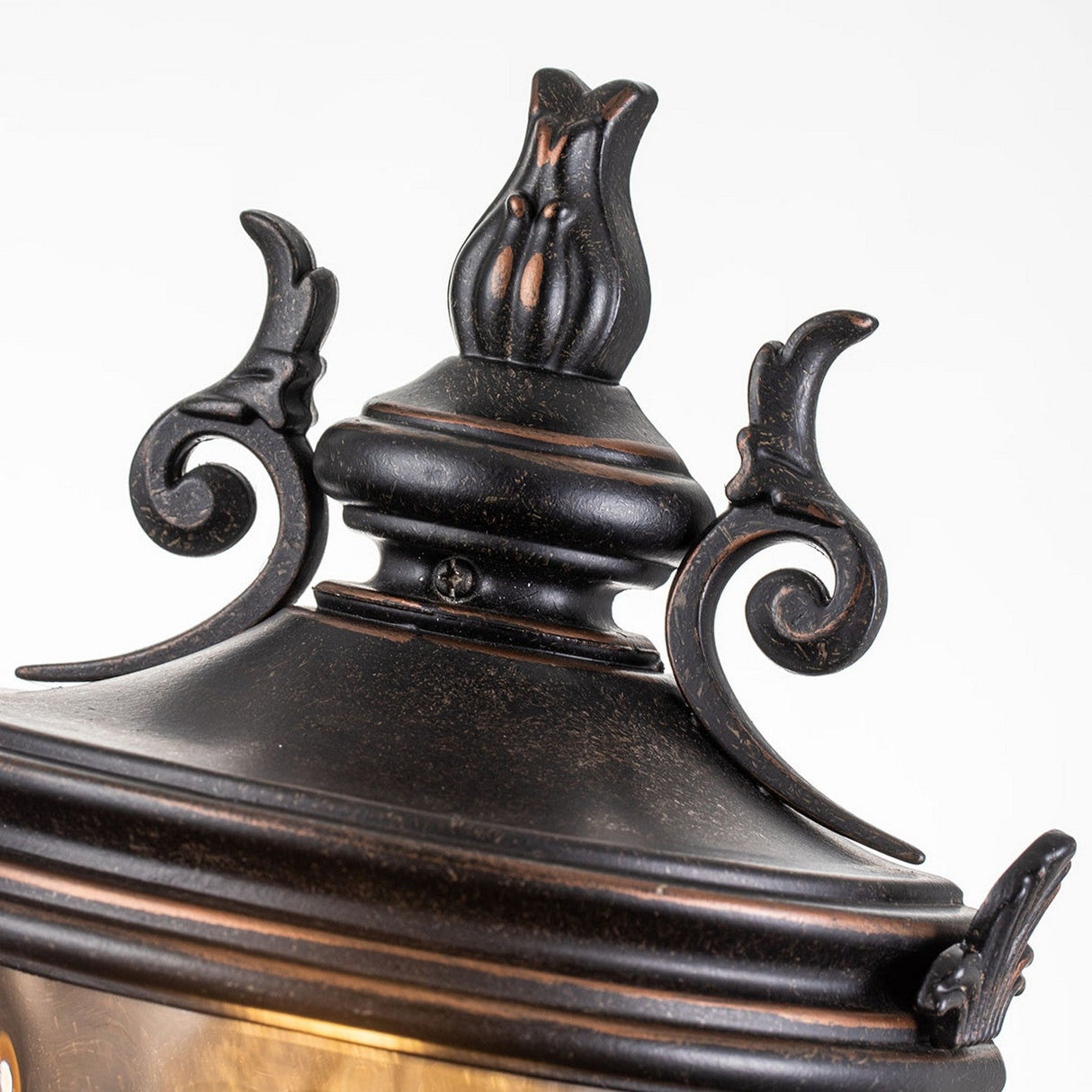 A close-up of the Baltimore 1 Light Outdoor Medium Half Lantern in weathered bronze showcases its ornate vintage-style design with intricate scrollwork, and amber-tinted glass accents that emit a warm glow against a light background.