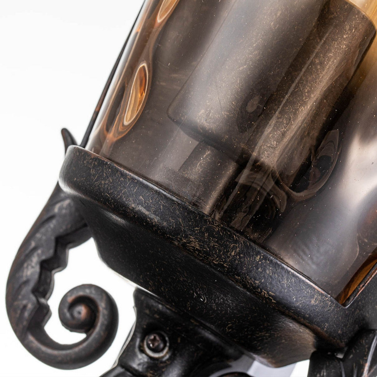 Close-up of the Baltimore 1 Light Outdoor Medium Half Lantern in weathered bronze. The black metal showcases intricate scrollwork, resembling a vintage lantern. The amber-tinted glass has a slightly frosted, antique appearance, with light reflecting subtly off its curved surfaces.