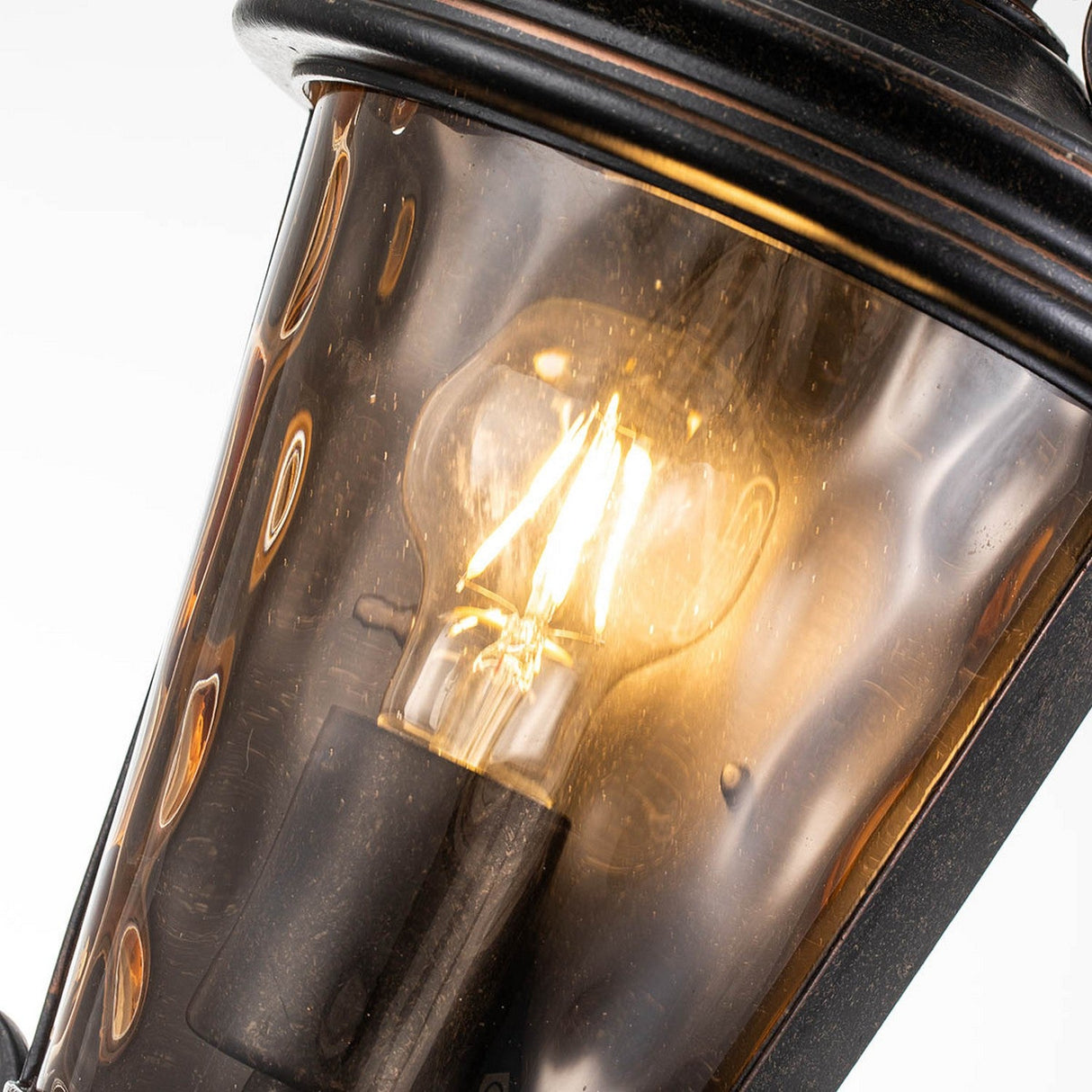 Close-up of a glowing light bulb inside the Baltimore 1 Light Outdoor Medium Half Lantern, featuring amber-tinted glass and a weathered bronze finish. The lantern's dark, polished metal frame creates a warm, inviting ambiance.