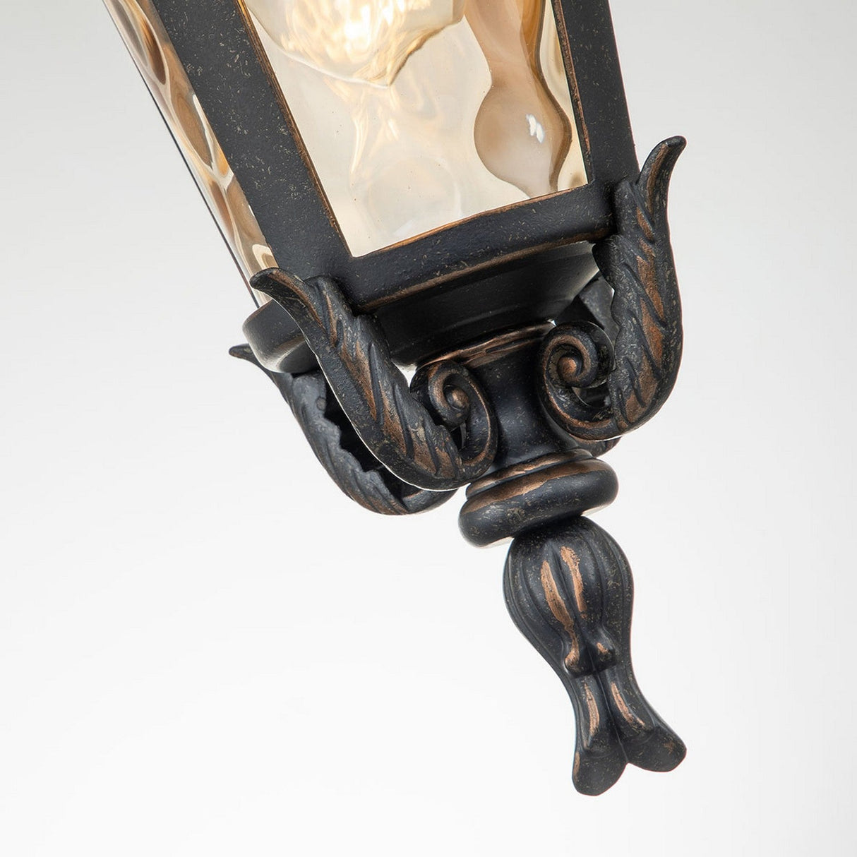 A close-up of the Baltimore 1 Light Outdoor Medium Chain Pendant in weathered bronze, featuring ornate scrollwork that exudes vintage charm. The elegant fixture encases a glowing light bulb, set against a plain white background to highlight its sophistication.