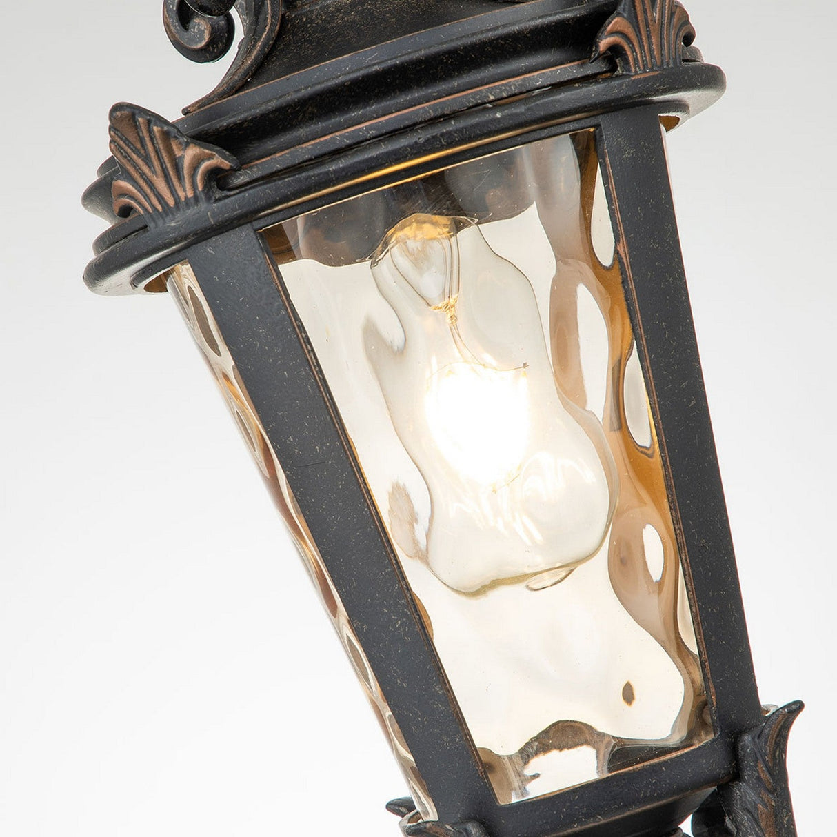 A detailed view of the Baltimore 1 Light Outdoor Medium Chain Pendant in a vintage style, featuring an incandescent bulb glowing within. This pendant boasts a weathered bronze metal frame adorned with decorative accents and textured glass panels, elegantly suspended from a medium chain.