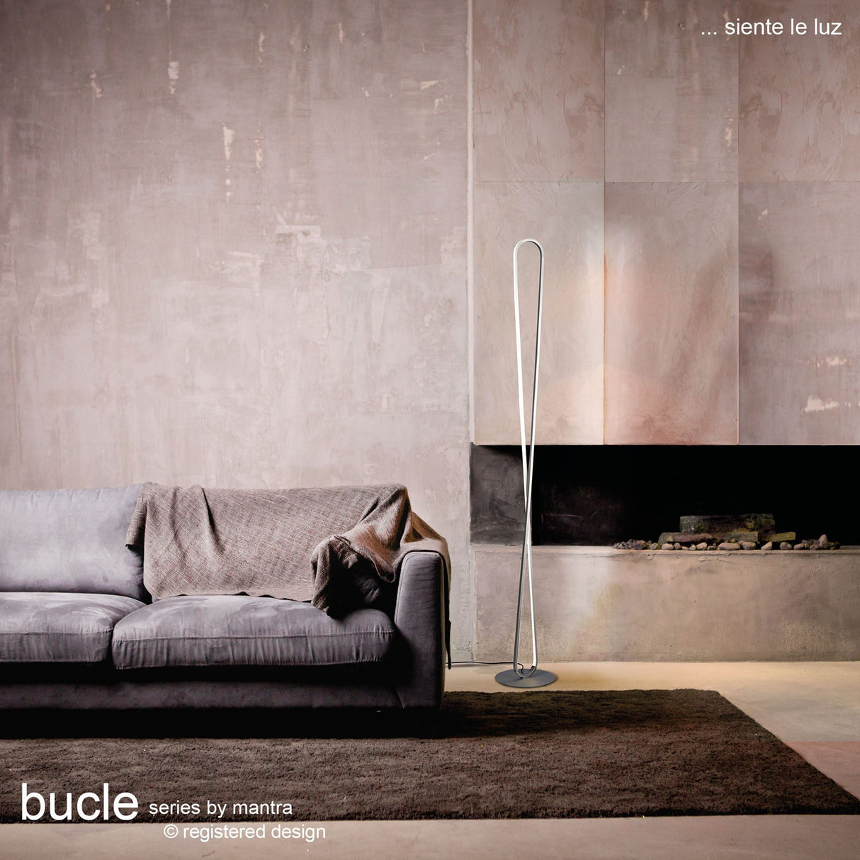 Modern living room with a gray sofa, minimalist fireplace, and the Bucle Dimmable LED Floor Lamp 50W 3000K in silver & chrome. Neutral decor adds to the contemporary aesthetic. “Bucle series by mantra” is mentioned in the image text.