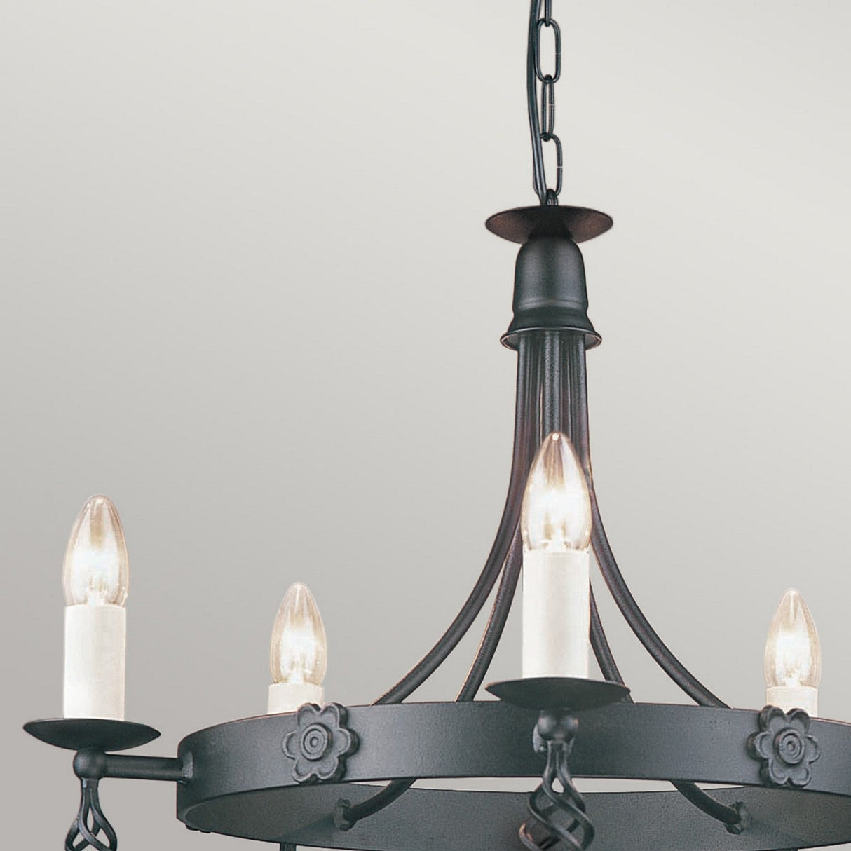 The Belfry 5 Light Chandelier - Black gracefully suspends from a chain, showcasing a black wrought iron structure and candle-shaped bulbs. Its traditional artistry is highlighted by floral accents that adorn the circular frame, set against a grey background, displaying exquisite hand-crafted detail.