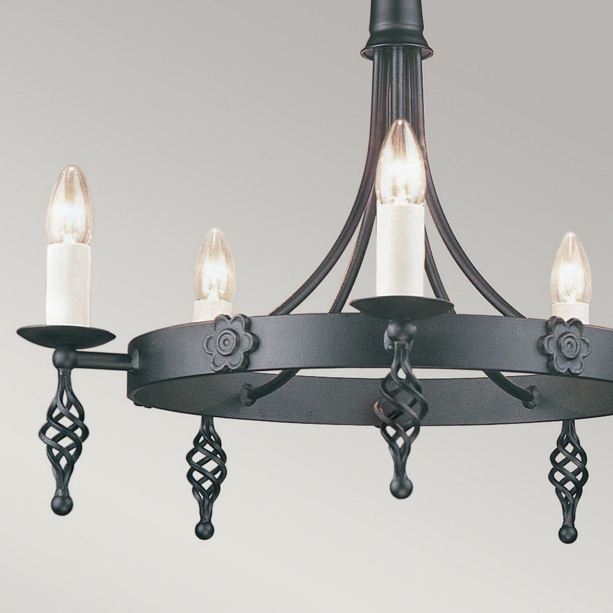 The Belfry 5 Light Chandelier - Black, a stunning example of traditional craftsmanship, showcases a black wrought iron frame and is adorned with four candle-shaped bulbs nestled in elaborately designed holders. This sophisticated and timeless piece is meticulously hand-crafted against a simple light gray backdrop.