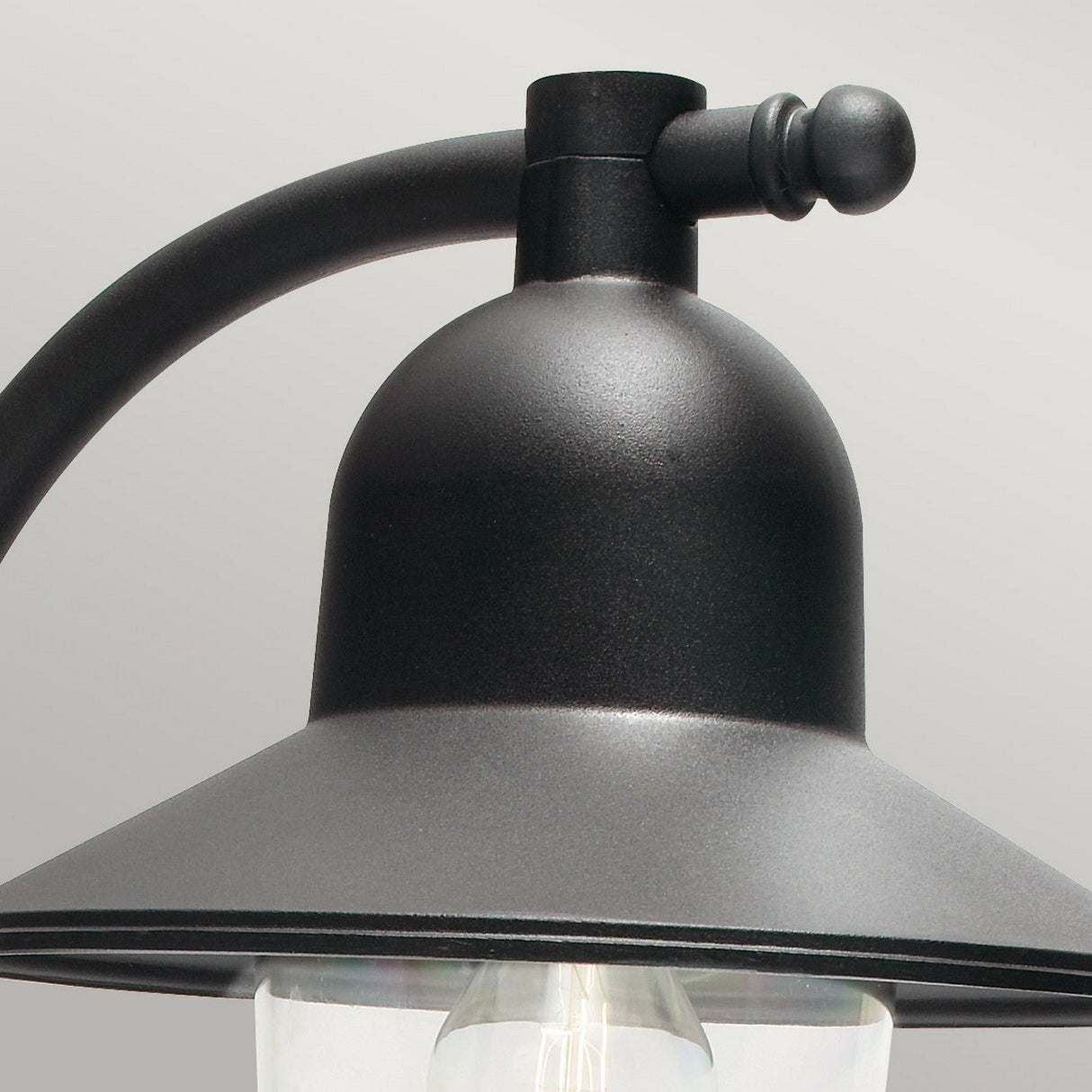 Close-up of the Como Outdoor 1 Light Wall Lantern in black, showcasing its curved arm and bell-shaped top. This wall lantern has a clear glass shade and a matte finish, set against a plain gray background.