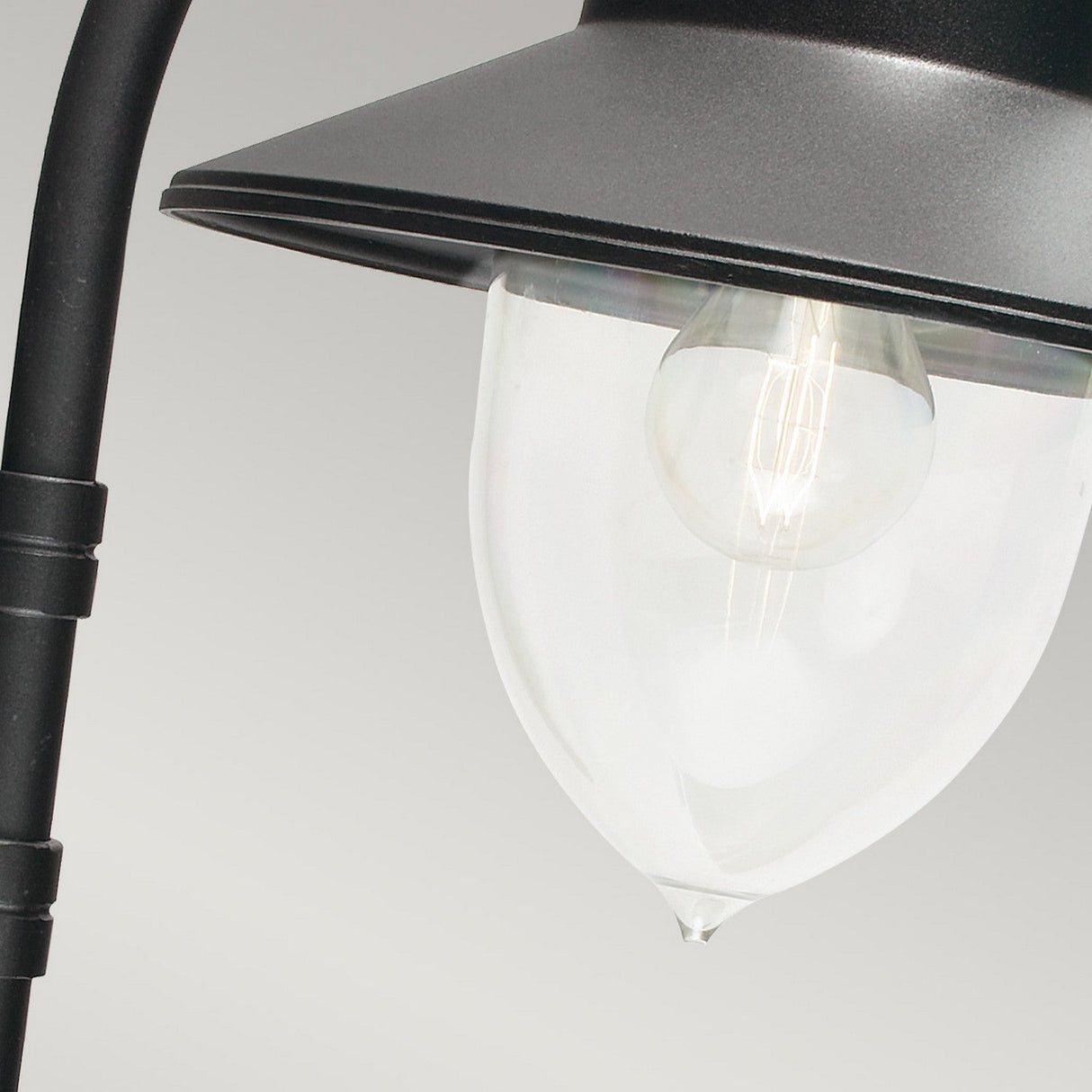 Close-up of the Como Outdoor 1 Light Wall Lantern in black, featuring a transparent glass bulb housing. The cast aluminum lantern is mounted on a black pole, highlighting the visible light bulb within. The background is a plain, light gray.