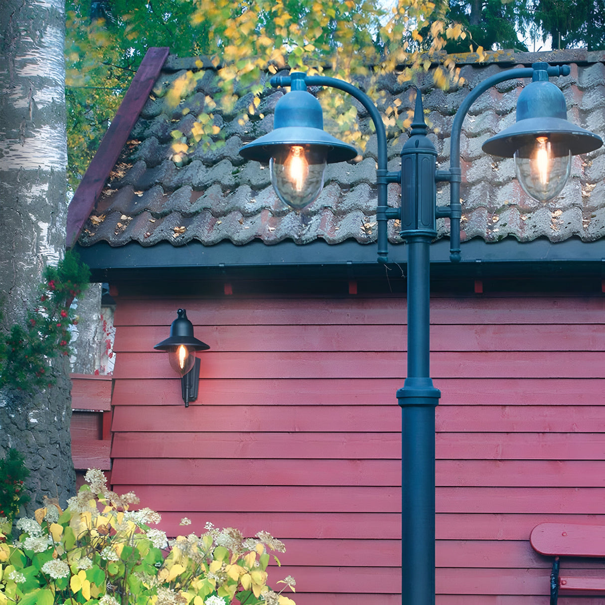 A rustic red wooden shed with a tiled roof stands in a garden setting. In the foreground, two Como Outdoor 1 Light Wall Lanterns - Black illuminate the area with a warm glow. The surrounding foliage exhibits autumnal hues, with leaves in shades of yellow and green.