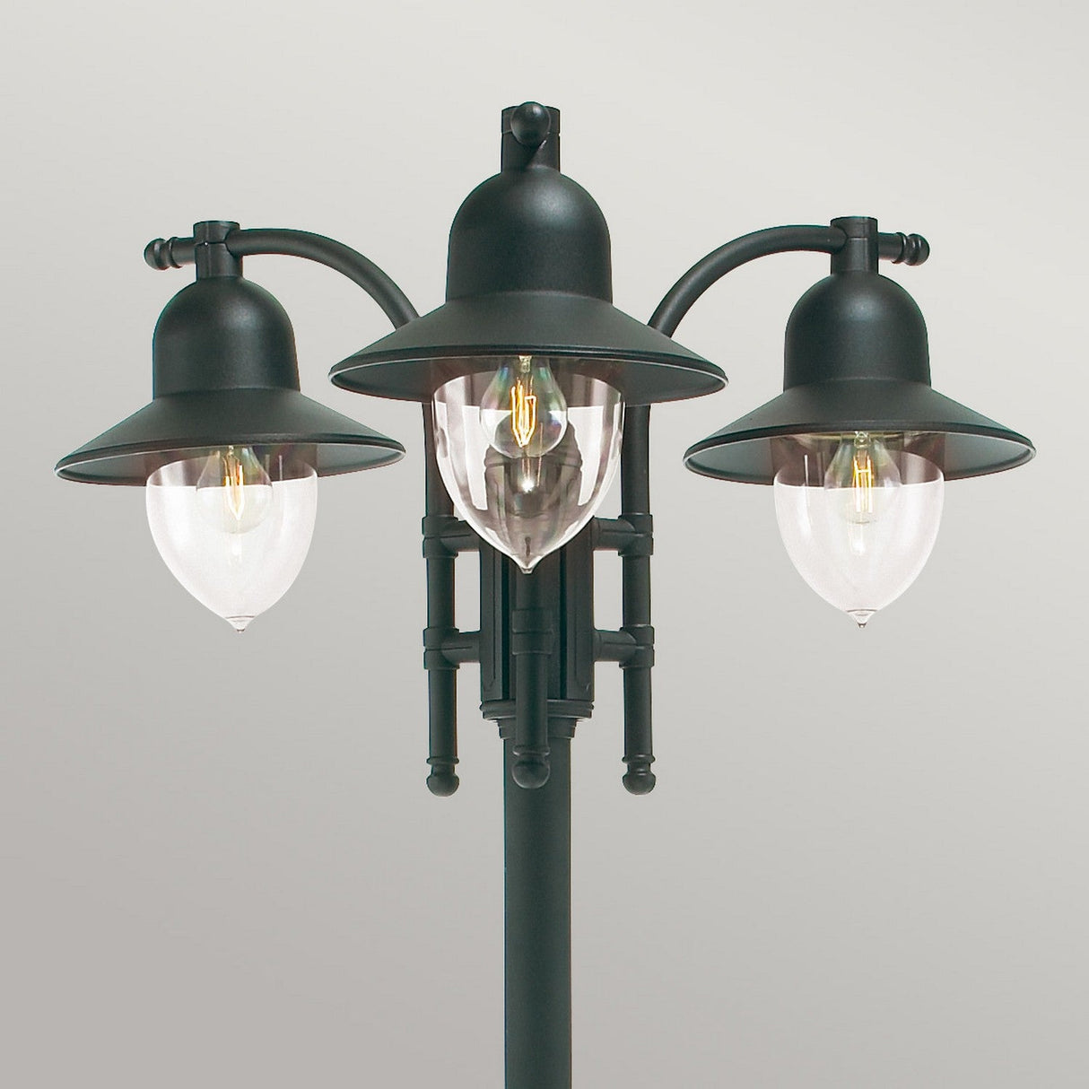 The Como 3 Light Triple Head Post - Black, designed for durable outdoor lighting with its IP54 rating, boasts black metal fixtures and clear glass shades that elegantly display an exposed filament bulb against a neutral backdrop.