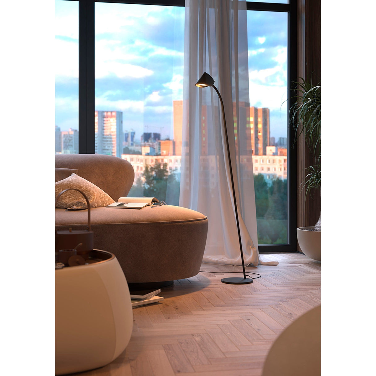 A cozy living room corner features a curved beige sofa with a soft throw, pillow, and open book. Ambient light comes from the Capuccina LED Floor Lamp 8.5W 3000K - Black, while a large window reveals a cityscape at sunset.
