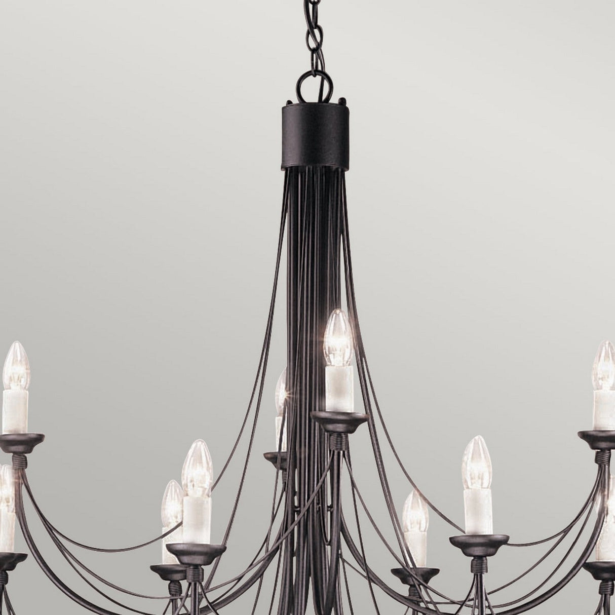 The Carisbrooke 12 Light Chandelier in Black features a Gothic design with multiple curved arms, each adorned with candle-shaped light bulbs. Its elegant and minimalist style is complemented by exposed wiring, adding a modern touch against the simple, off-white wall.