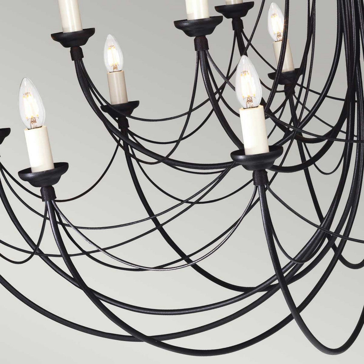 The Carisbrooke 18 Light Chandelier - Black delivers a modern, Gothic-inspired aesthetic with its close-up view revealing candelabra-style arms crafted from black metal and adorned with white candle-shaped bulbs. Set against a plain backdrop, the chandelier's elegant and intricate design is characterized by its curving lines and sophisticated light fixtures.